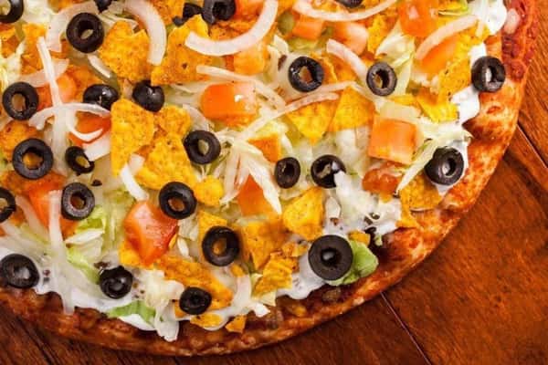 Taco pizza topped with shredded lettuce, tomatoes, onions, black olives, and crushed nacho chips on a wooden table.