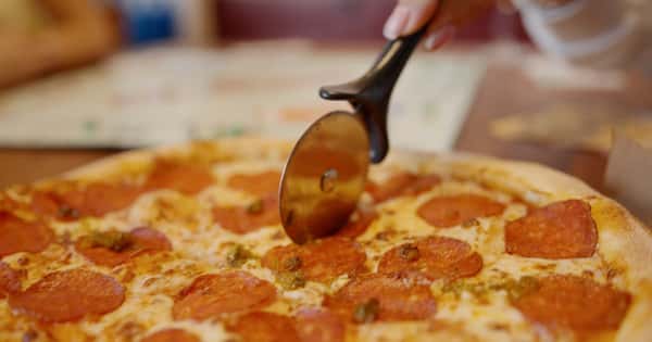 Hand slicing a hot pepperoni pizza with a pizza cutter, featuring melted cheese and jalapeño slices on a golden crust.