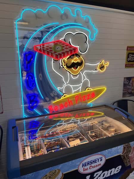 Neon Beach Pizza sign featuring a surfing chef with a pizza box above a Hershey's ice cream freezer stocked with frozen treats.