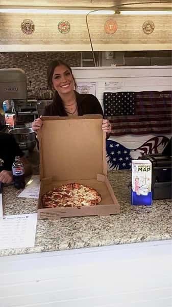 Smiling woman behind a counter holding an open pizza box with a fresh pepperoni pizza inside, American flag decor in background.