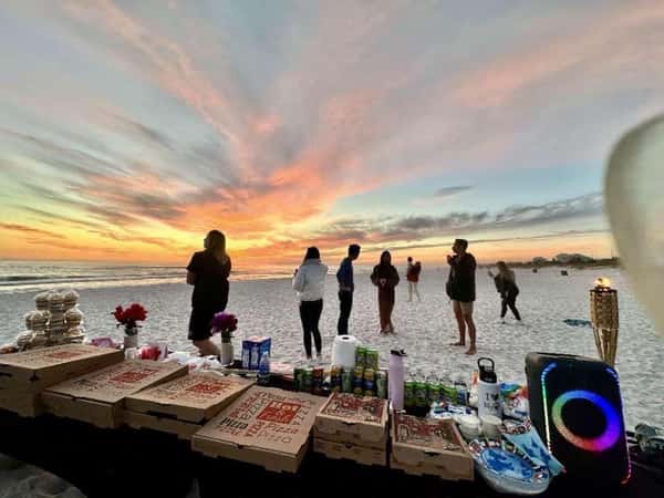 Pizza boxes, drinks, and snacks on a beach table at sunset with people gathered and a colorful sky in the background.