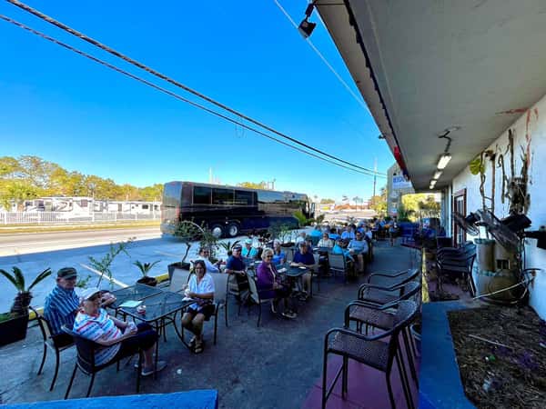 Customers dining on the shaded patio outside Beach Pizza, with potted palms, street view, and a tour bus parked nearby.