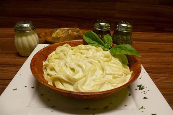 Fettuccini Alfredo in a bowl with basil leaves on top. Parmesan cheese, spices and garlic knots on the side.