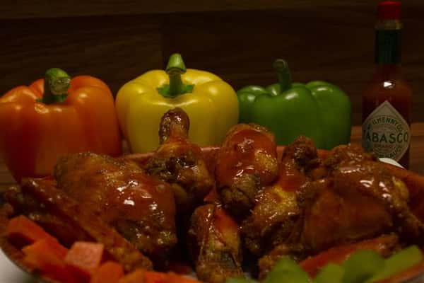 Close up of Chicken wings in a bowl with a bottle of Tabasco sauce and assorted bell peppers on the side.