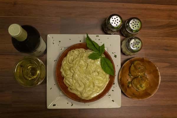 Top view of Fettuccini Alfredo in a bowl with basil leaves on top. Parmesan cheese, spices and garlic knots on the side. Bottle of Terre Di Chieti Chardonnay poured into a wine glass.