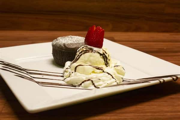 Side view of Chocolate Souffle on a square plate with a scoop of vanilla ice cream on the side. Ice cream topped with a strawberry and chocolate syrup drizzle.