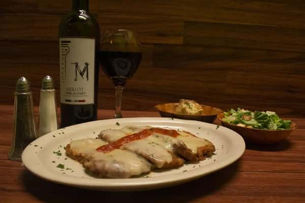 Chicken Parmigiana topped with melted mozzarella cheese and a touch of tomato sauce. Garlic knots and small Caesar salad on the side. Bottle of Terre Di Chieti Merlot poured into a wine glass.