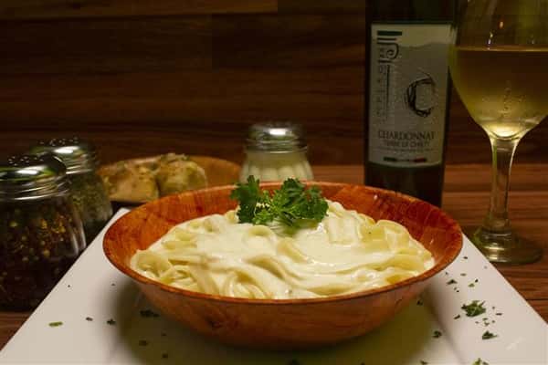 Fettuccini Alfredo in a bowl with basil leaves on top. Parmesan cheese, spices and garlic knots on the side. Bottle of Terre Di Chieti Chardonnay poured into a wine glass.