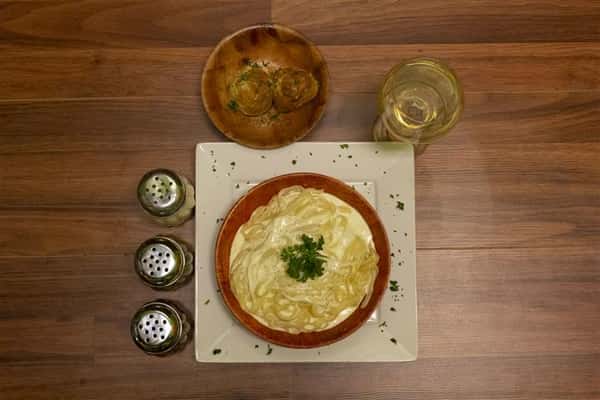 Top view of Fettuccini Alfredo in a bowl with basil leaves. Glass of white wine, spices and bowl of garlic knots on the side.