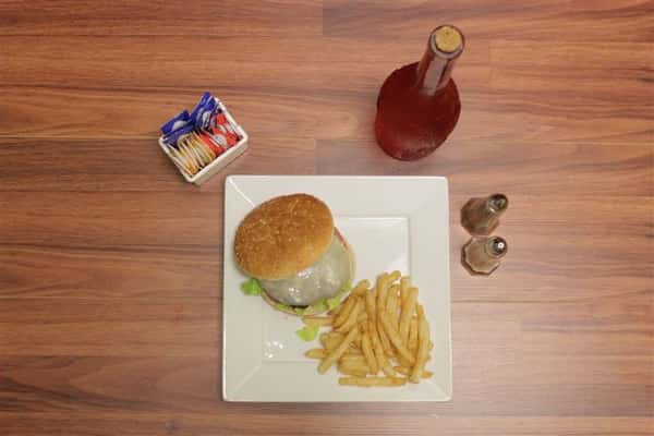 Top view of Cheeseburger topped with Swiss cheesewith lettuce, tomato and onion. French fries, salt, pepper, sauces and unopened bottle of rose on the side.