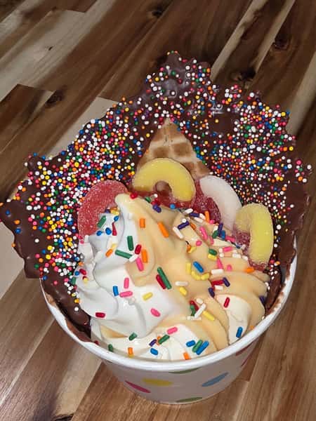 Over-the-top frozen yogurt sundae in a chocolate-dipped waffle bowl with rainbow sprinkles, gummy candy slices, and creamy soft serve – colorful specialty dessert.