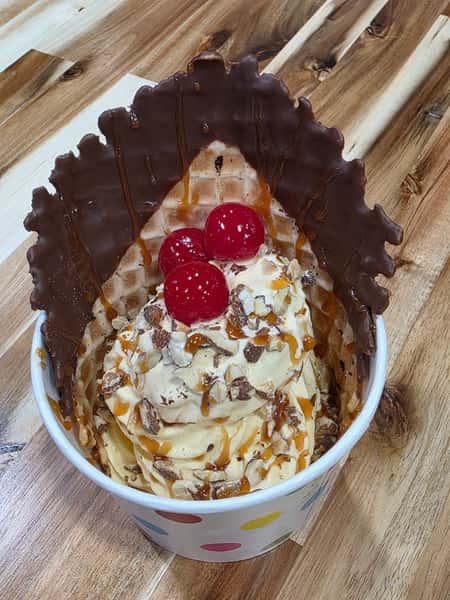 Caramel pecan frozen yogurt sundae with chocolate-dipped waffle bowl and maraschino cherries – decadent soft serve dessert with caramel drizzle and nuts.