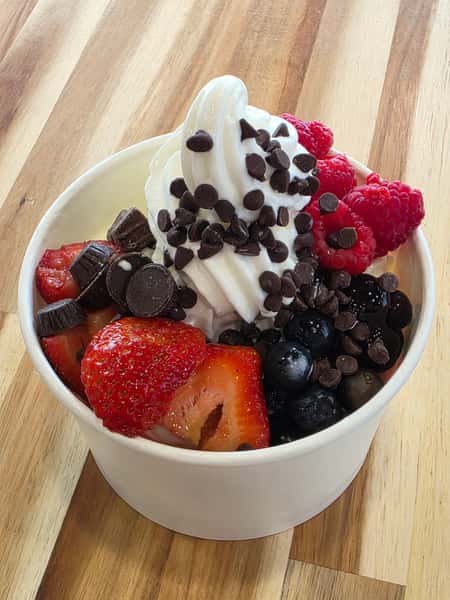 Vanilla frozen yogurt with fresh strawberries, raspberries, blueberries, mini chocolate chips, and peanut butter cups – fruit and chocolate froyo bowl on wooden counter.