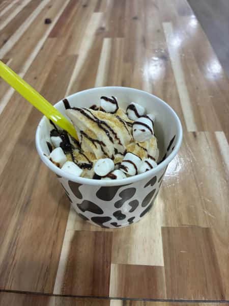 Vanilla soft serve with mini marshmallows, graham cracker crumble, and chocolate syrup drizzle in a cow-print cup – s'mores-inspired frozen yogurt dessert served on a rustic wooden counter.