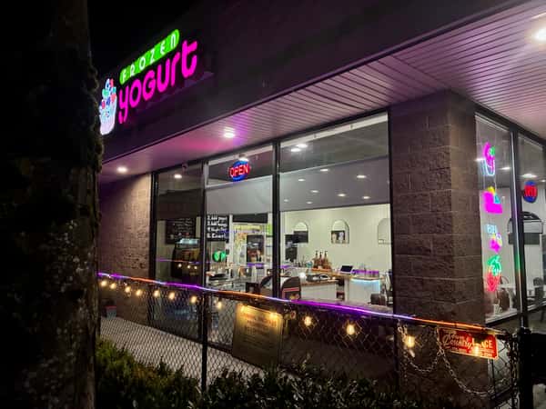 Exterior of a frozen yogurt shop at night with bright neon "Frozen Yogurt" signage, glowing open sign, and colorful window lights – modern dessert café storefront with inviting ambiance, perfect for a local ice cream or froyo business listing.
