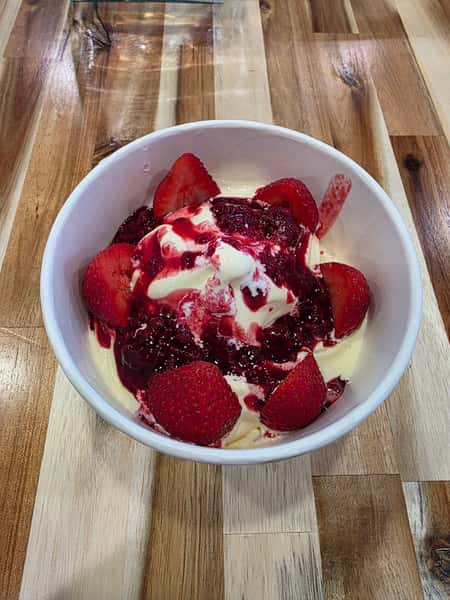 Strawberry cheesecake dessert topped with fresh sliced strawberries and mixed berry compote in a white bowl, served on a rustic wooden table – creamy New York-style cheesecake with vibrant red berry sauce, perfect for a modern café or dessert restaurant menu.