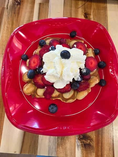 Golden bubble waffle topped with fresh strawberries, blueberries, and a generous dollop of whipped cream on a red plate – fruity dessert waffle perfect for brunch or a sweet café treat.