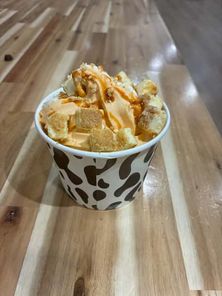Pumpkin spice soft serve topped with toasted marshmallow cubes and caramel drizzle in a cow-print cup – seasonal fall frozen yogurt flavor ideal for autumn dessert specials.