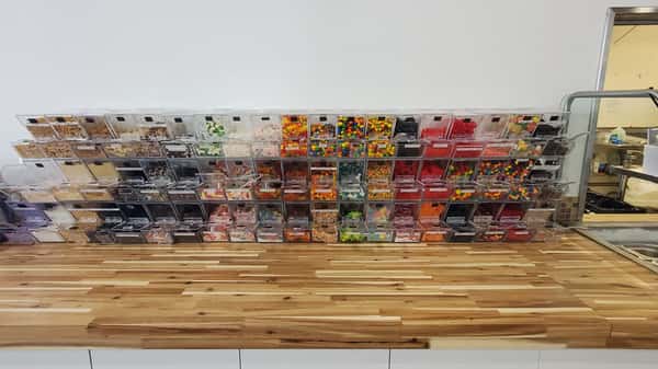 Self-serve frozen yogurt topping station with rows of candy, sprinkles, and nuts in clear bins – customizable froyo bar inside a modern dessert shop.
