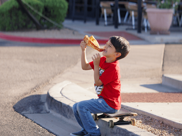 Boy sitting on a skateboard eating a pizza slice