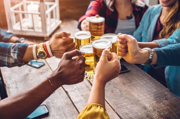 Hands raise glasses of beer in a toast, showcasing diverse skin tones and accessories, against a rustic wooden table, capturing a moment of celebration and camaraderie.