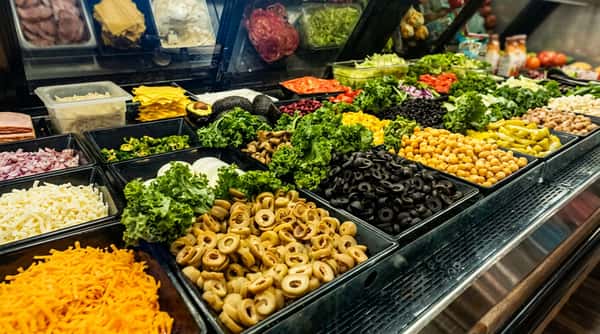 Fresh salad bar with colorful toppings including lettuce, olives, chickpeas, shredded cheese, peppers, onions, and vegetables displayed in a restaurant deli counter for custom salads and healthy dining.