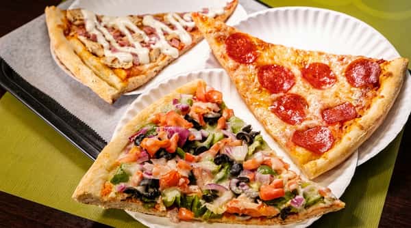 Assorted pizza slices featuring pepperoni, vegetable, and chicken specialty toppings, served fresh on paper plates in a casual pizzeria.