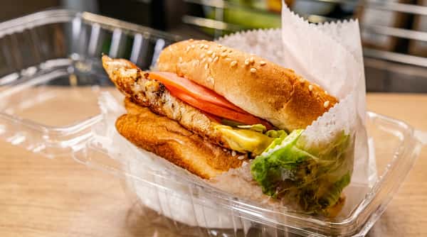 Grilled chicken sandwich with lettuce, tomato, and sauce on a sesame seed bun, in a clear plastic container.