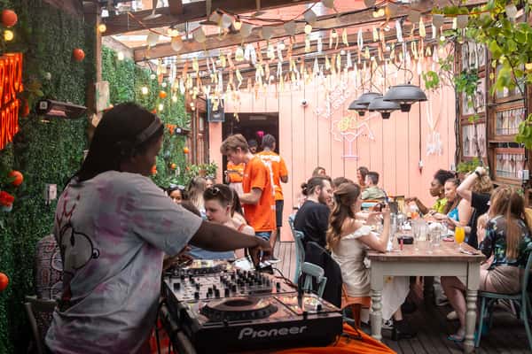 Lively outdoor restaurant patio with guests dining at communal tables, a DJ mixing music in the foreground, string lights overhead, and colorful cocktails creating a vibrant social dining atmosphere.