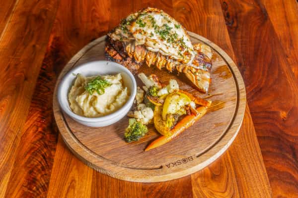 Grilled lobster tail with herb butter, mashed potatoes, and roasted seasonal vegetables served on a wooden board.