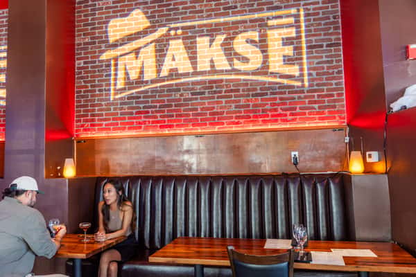 Makse restaurant dining room with exposed brick, red accent lighting, leather booth seating, and guests enjoying drinks.