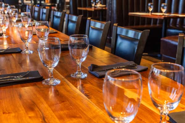 Elegant restaurant dining area with a long wooden table set with water glasses, black napkins, and dark leather chairs.