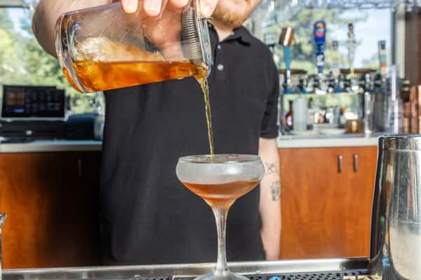 Bartender pouring a freshly mixed craft cocktail into a coupe glass at the restaurant bar.