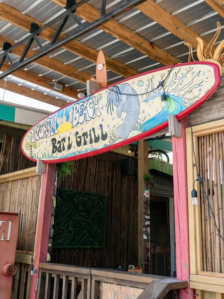 Entrance to North Beach Bar & Grill featuring a colorful surfboard sign with a manatee illustration, under a wooden patio roof.