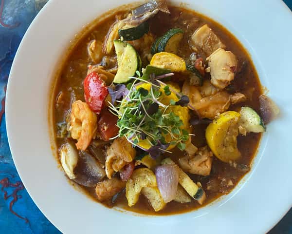 Seafood and vegetable stew with shrimp, fish, zucchini, squash, and tomatoes in a rich broth, topped with microgreens.