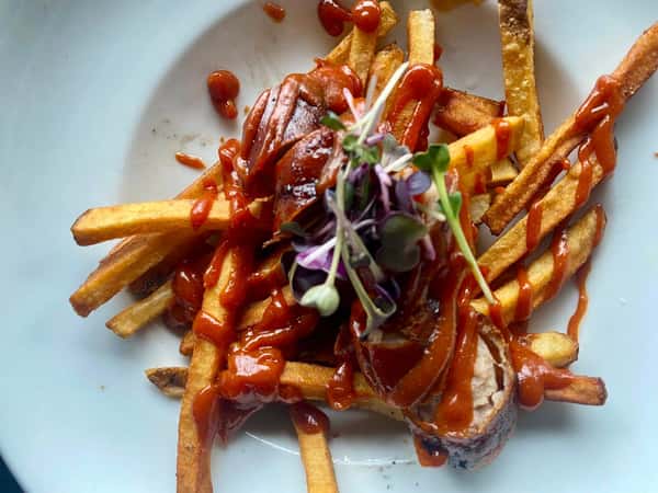 Crispy French fries topped with sliced sausage, drizzled ketchup, and fresh microgreens on a white plate.