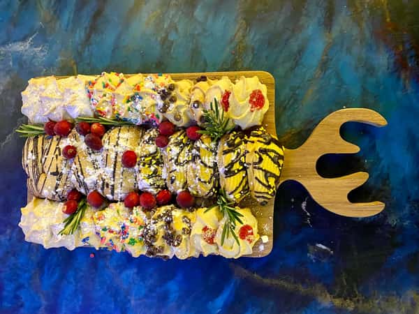 Assorted cream-filled cannoli topped with powdered sugar, sprinkles, cherries, and chocolate drizzle on a fish-shaped wooden board.