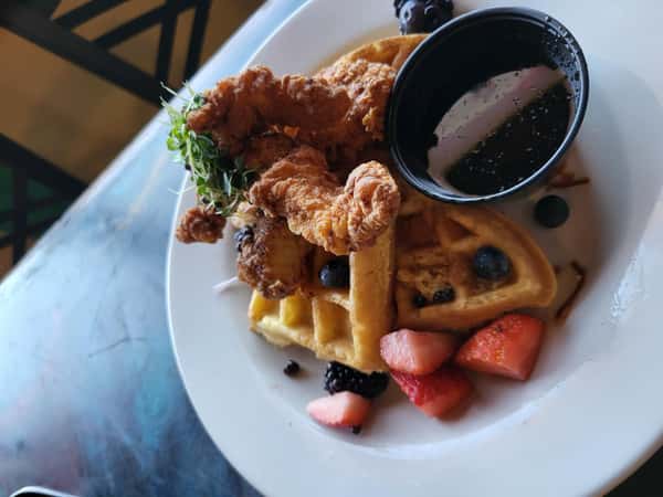 Fried chicken and waffles served with fresh berries, microgreens, and a side of syrup on a white plate.