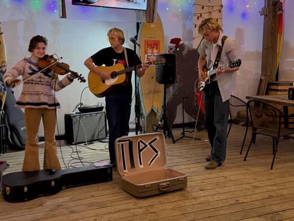 Three-piece band performing indoors with a violinist, acoustic guitarist, and electric guitarist, with a tip suitcase open in front.