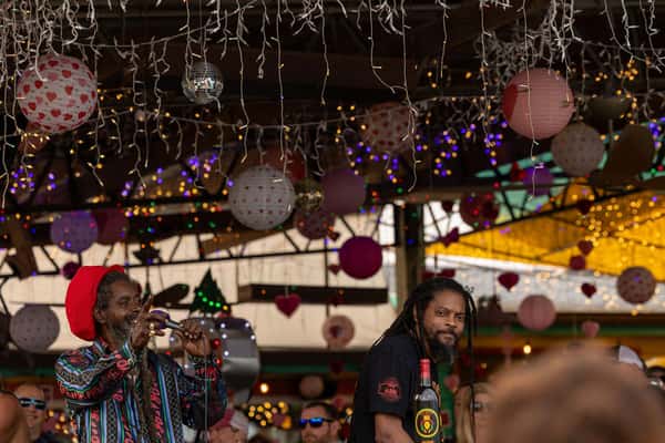 This photo captures a lively moment likely at a performance or event at North Beach Bar and Grill. One man sings or speaks into a microphone while another stands nearby, both with Rastafarian-inspired style. The background is filled with festive hanging decorations, string lights, and hearts—suggesting a celebration or themed party, possibly Valentine's Day. The warm, colorful ambiance and vibrant energy reflect a fun, island-style atmosphere.