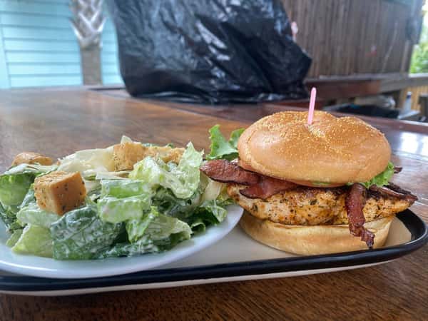 Grilled chicken sandwich with bacon, lettuce, and tomato on a bun, served with a side Caesar salad topped with croutons.