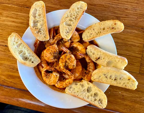 Spicy seasoned shrimp served in sauce with toasted baguette slices arranged around the edge of a white plate.