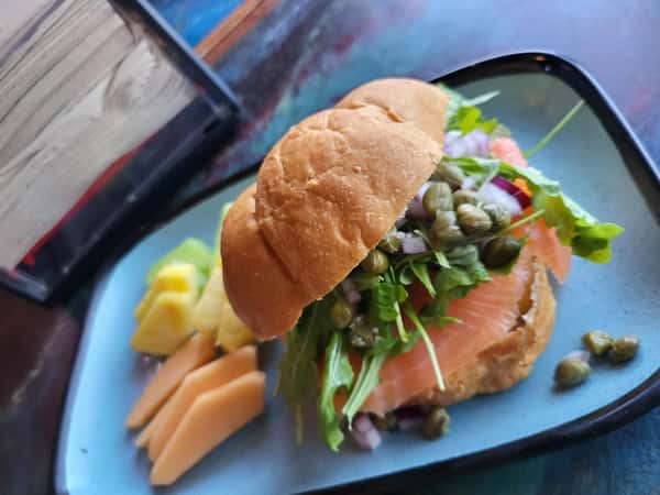 Smoked salmon sandwich with capers, greens, and red onions on a bun, served with fresh pineapple and cantaloupe slices.