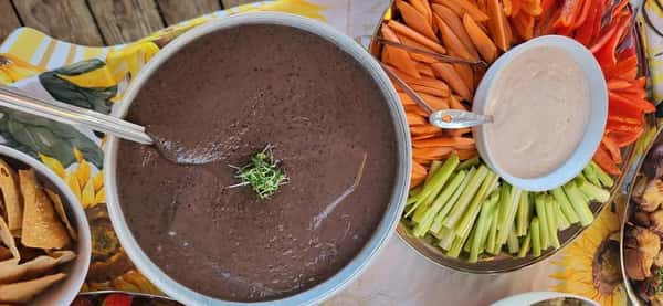 Bowl of black bean soup garnished with microgreens, surrounded by pita chips, fresh veggie sticks, and a bowl of dipping sauce.
