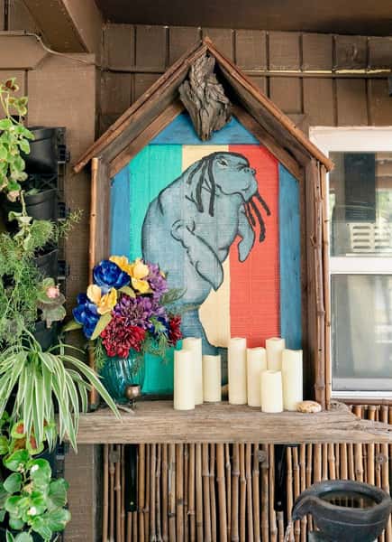 Rustic altar-style display featuring a painted manatee with dreadlocks on a tri-color wood background, surrounded by candles and flowers.