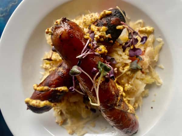 Grilled sausages served over sauerkraut, topped with whole grain mustard and microgreens on a white plate.