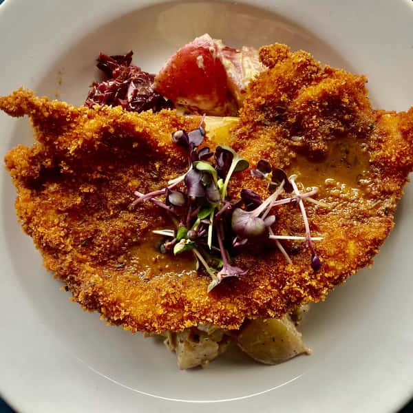 Crispy breaded pork schnitzel topped with microgreens, served with potato salad and red cabbage on a white plate.