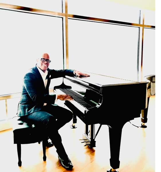 Cornelius Herring sits at a grand piano in a well-lit room. He is wearing a dark suit, white shirt, and glasses, with a friendly expression and a slight smile. The background is brightly illuminated, likely from large windows behind him, creating a high contrast between the subject and the surroundings. The floor is wooden, and the piano bench and piano have a polished black finish.