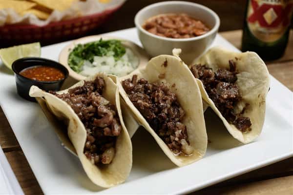 Three soft tacos with steak and onions on a white plate with a cup of salsa, onions, and beans