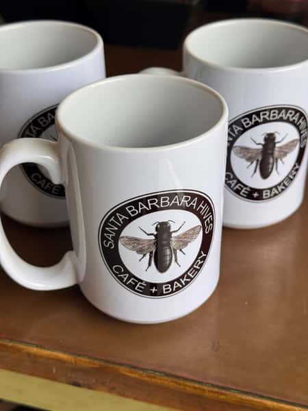 White ceramic mugs with the Santa Barbara Hives Café + Bakery bee logo printed on them.